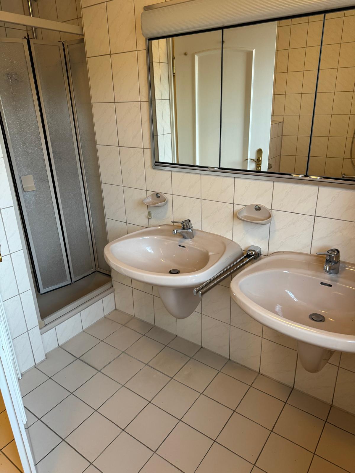 Altes Bad mit beigen Fliesen, Doppelwaschbecken und Duschkabine vor der Sanierung in Augsburg